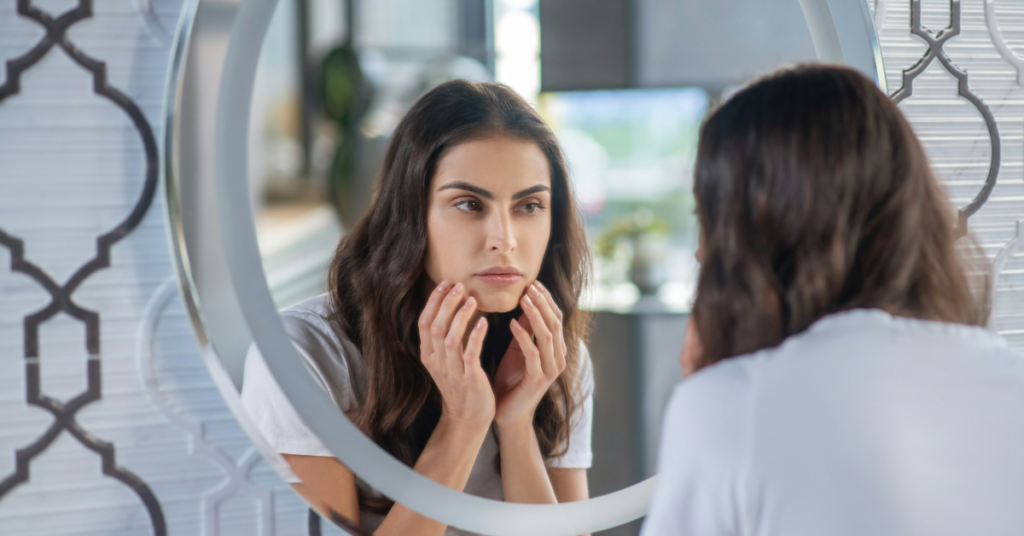 Beautiful young woman looking in mirror touching her chin and checking for cortisol face