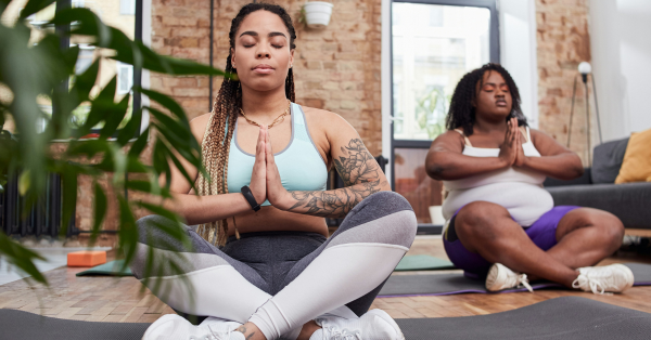 Two Black women sitting in a yoga pose on mats in a small room