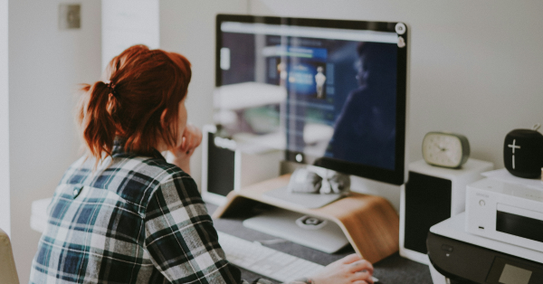 Young woman with red hair wearing a plaid flannel shirt sitting at computer desk and working 