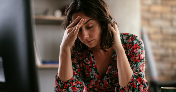 Stressed young woman with short brown hair wearing a floral blouse sitting at desk with her head in her hands and eyes closed