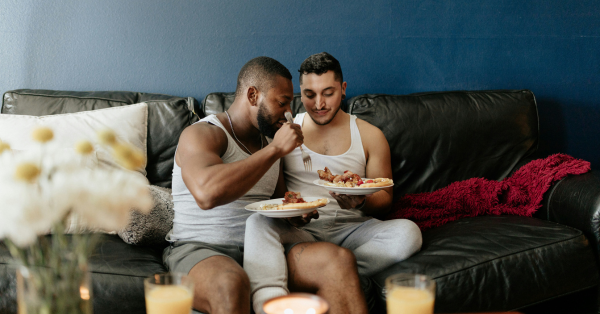 Two queer men sitting on couch together having a date night dinner at home