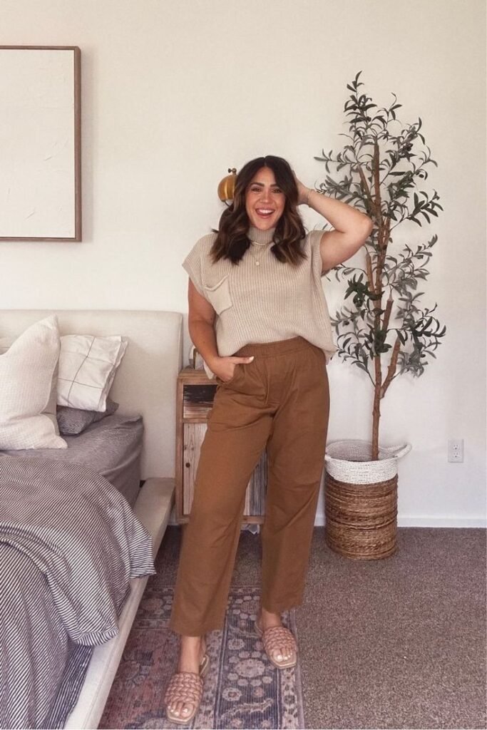 Brunette woman wearing sleeveless sweater top and brown pants with slip on sandals for a cute fall transition outfit